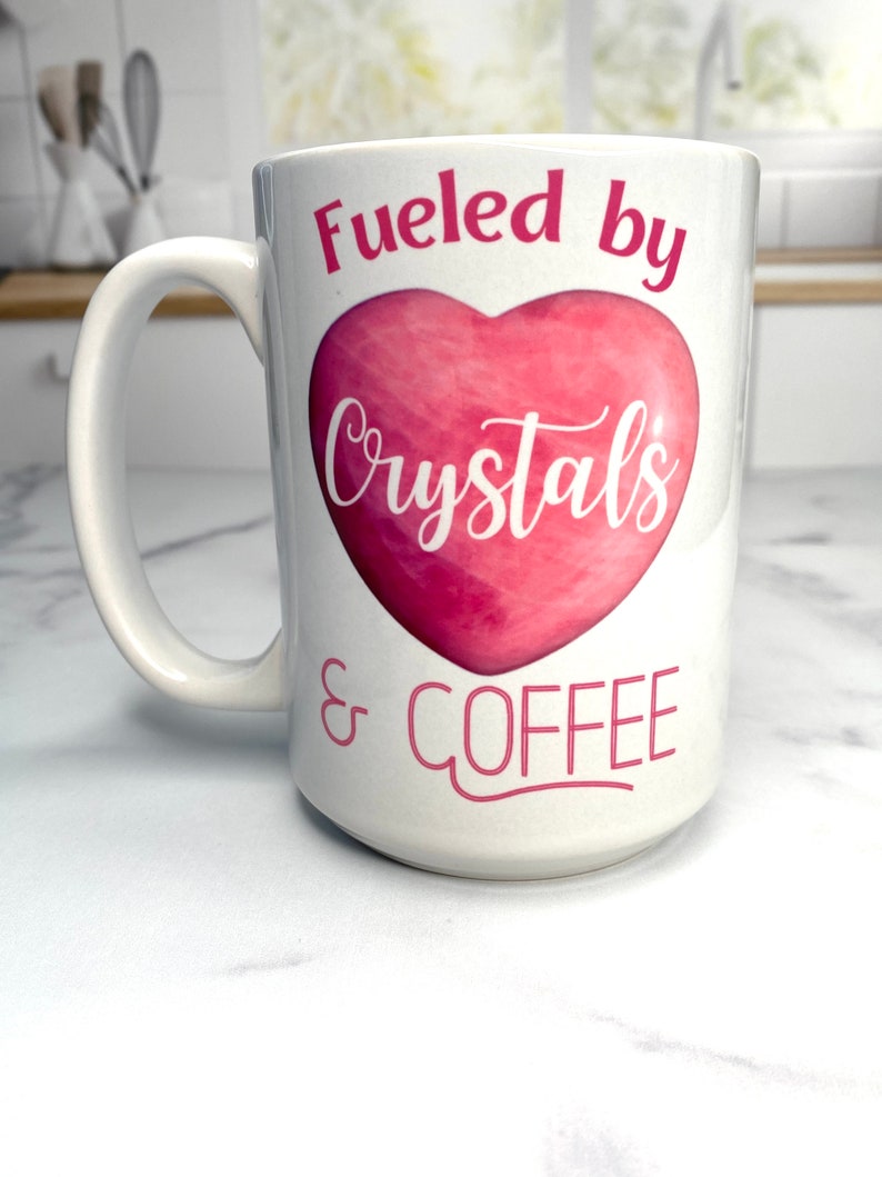 May include: White ceramic coffee mug with a pink heart and the text "Fueled by Crystals & Coffee".