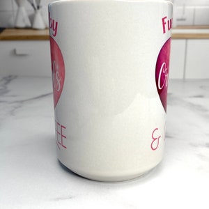 May include: White ceramic mug with a pink heart design. The text on the mug reads "Fueled by Coffee & Crazy".