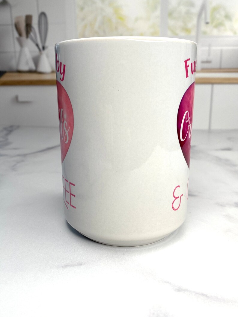 May include: White ceramic mug with a pink heart design. The text on the mug reads "Fueled by Coffee & Crazy".
