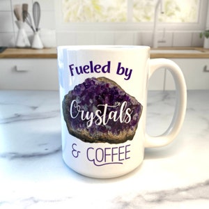 Peut inclure: Une tasse &agrave; caf&eacute; en c&eacute;ramique blanche avec un motif de cristal d'am&eacute;thyste violet et le texte "Fueled by Crystals & Coffee".