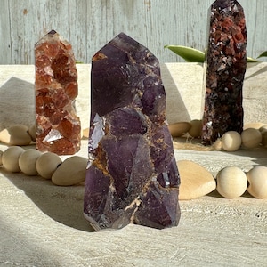May include: Three amethyst crystal points with inclusions of other minerals. The crystals are a deep purple color and have a rough, natural texture. The crystals are arranged on a white wooden surface.