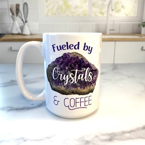 Peut inclure: Une tasse &agrave; caf&eacute; en c&eacute;ramique blanche avec un motif de cristal d'am&eacute;thyste violet et le texte "Fueled by Crystals & Coffee".