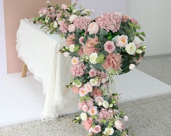 Puede incluir: Una larga guirnalda de flores artificiales en tonos rosa y blanco, con vegetación, se coloca sobre una mesa blanca. La guirnalda incluye rosas, hortensias y otros elementos florales.