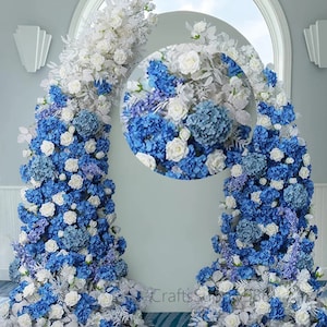 Op de afbeelding: Een bruiloftsboog versierd met witte en blauwe kunstbloemen. De boog bestaat uit twee aparte delen, elk met een mix van witte rozen en blauwe hortensia's.