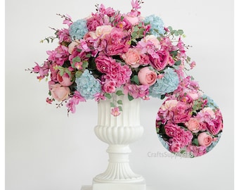 May include: A white ceramic urn vase filled with a large arrangement of pink, peach, and light blue artificial flowers. The flowers are arranged in a cascading style, with some flowers hanging over the edge of the vase.