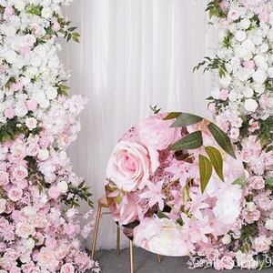 Op de afbeelding: Een roze en witte bloemenachtergrond met een ronde, roze bloemenarrangement in het midden. De achtergrond is gemaakt van kunstbloemen en groen.