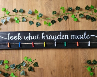 May include: A black wooden sign with the text "look what brayden made" in white cursive lettering. A string with colorful clothespins is attached to the sign.