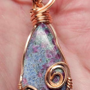 May include: A teardrop-shaped pendant with a blue, green, and pink gemstone wrapped in copper wire. The pendant is attached to a black cord.