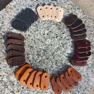 May include: A collection of leather strap loops in various colours, including brown, tan, black, and cream. The loops are arranged in a circle on a textured surface.