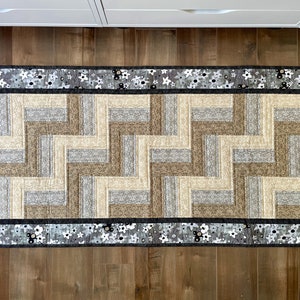 May include: A gray and beige patterned rug with a black and white floral border. The rug has a geometric design with a zig-zag pattern.