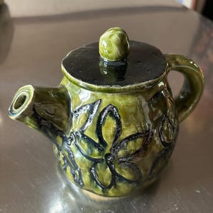 May include: A green ceramic teapot with a black lid and a floral design. The teapot has a rounded body and a spout on the side. The floral design is in black and is raised from the surface of the teapot.