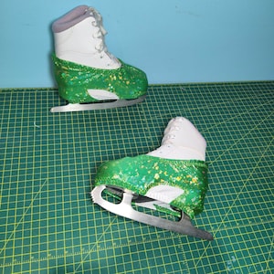 May include: Two white figure skates with green glitter on the boot. The skates are on a green mat.