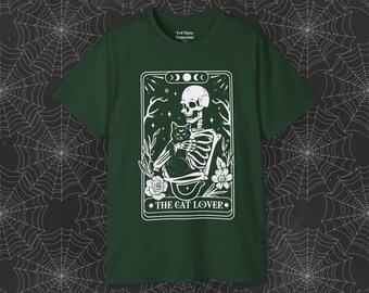 Puede incluir: Camiseta verde con un gráfico blanco de un esqueleto sosteniendo un gato negro. El gráfico está en el estilo de una carta de tarot y dice "The Cat Lover" en la parte inferior.