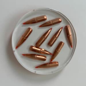 May include: A close-up of a clear round container with ten copper-colored bullets with red tips. The bullets are arranged in a circular pattern.