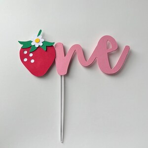 K&ouml;nnte beinhalten: Ein rosa Papier-Kuchentopper mit dem Wort "one" in Kursivschrift. Eine rote Papier-Erdbeere mit gr&uuml;nen Bl&auml;ttern und einer wei&szlig;en Bl&uuml;te ist an der linken Seite des Toppers befestigt.