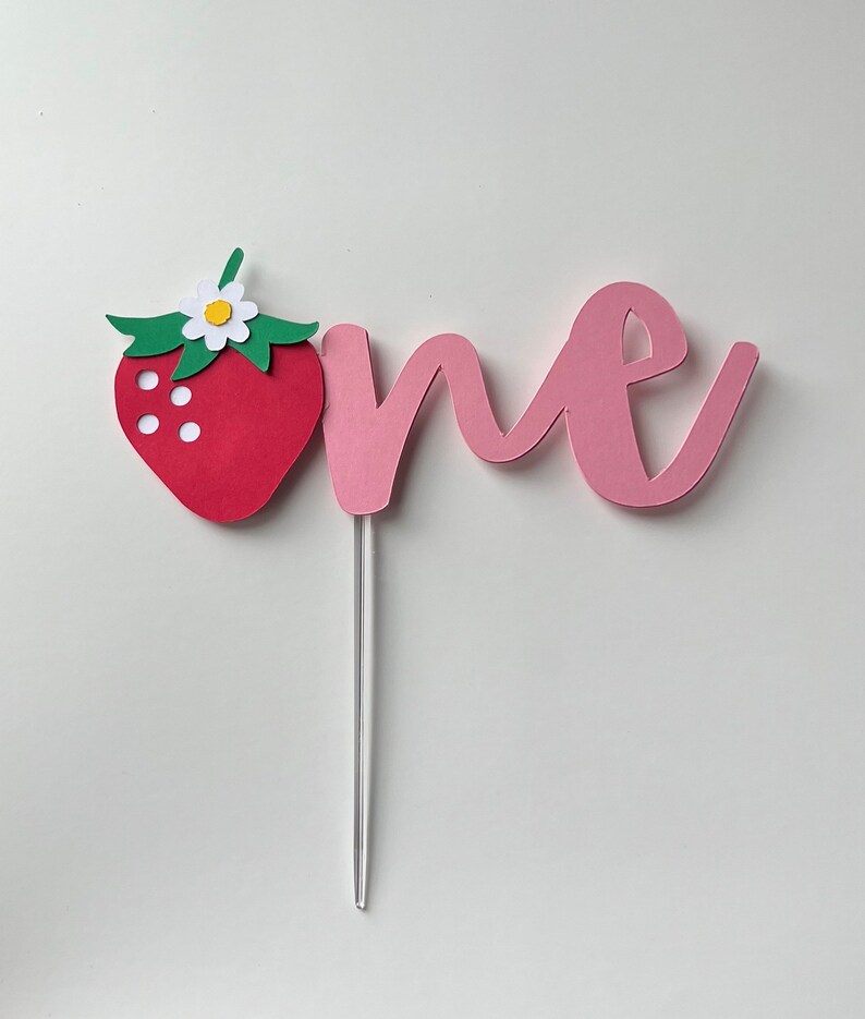 K&ouml;nnte beinhalten: Ein rosa Papier-Kuchentopper mit dem Wort "one" in Kursivschrift. Eine rote Papier-Erdbeere mit gr&uuml;nen Bl&auml;ttern und einer wei&szlig;en Bl&uuml;te ist an der linken Seite des Toppers befestigt.