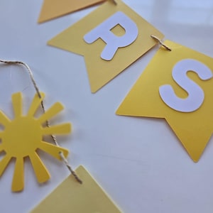 Puede incluir: Una pancarta de papel amarilla y blanca con la palabra "BRIGHTS" escrita en letras blancas. La pancarta est&aacute; enhebrada con un cordel y tiene un recorte de sol amarillo unido a ella.
