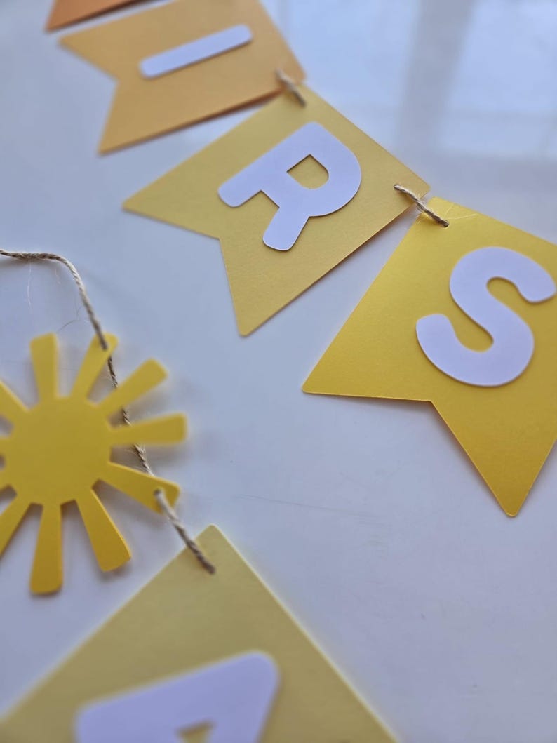 Puede incluir: Una pancarta de papel amarilla y blanca con la palabra "BRIGHTS" escrita en letras blancas. La pancarta est&aacute; enhebrada con un cordel y tiene un recorte de sol amarillo unido a ella.