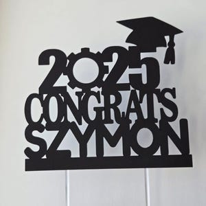 May include: Black graduation cake topper with the year 2023, the word "Congrats", and the name "Saymon".