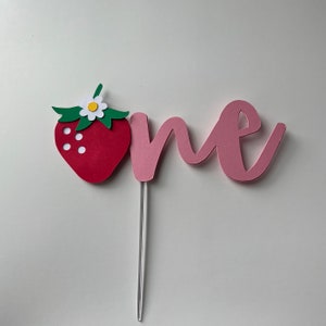 K&ouml;nnte beinhalten: Rosafarbener Papier-Kuchentopper mit dem Wort "one" und einer roten Papier-Erdbeere mit einer wei&szlig;en Bl&uuml;te und gr&uuml;nen Bl&auml;ttern.