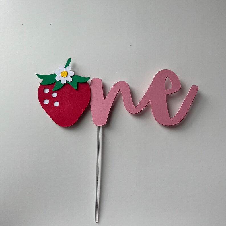 K&ouml;nnte beinhalten: Rosafarbener Papier-Kuchentopper mit dem Wort "one" und einer roten Papier-Erdbeere mit einer wei&szlig;en Bl&uuml;te und gr&uuml;nen Bl&auml;ttern.