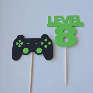 May include: Two green and black paper cake toppers. One topper is a video game controller and the other is a number 8 with the word "LEVEL" above it.
