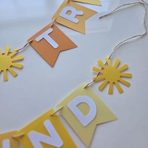Puede incluir: Una pancarta de papel amarilla y naranja con la palabra "SUNDAY" en letras blancas. La pancarta est&aacute; enhebrada con un cordel y tiene recortes de sol amarillos.