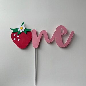 K&ouml;nnte beinhalten: Ein rosa Papier-Kuchentopper mit dem Wort "one" in kursivschrift. Eine rote Papier-Erdbeere mit gr&uuml;nen Bl&auml;ttern und einer wei&szlig;en Bl&uuml;te ist an der linken Seite des Toppers befestigt.