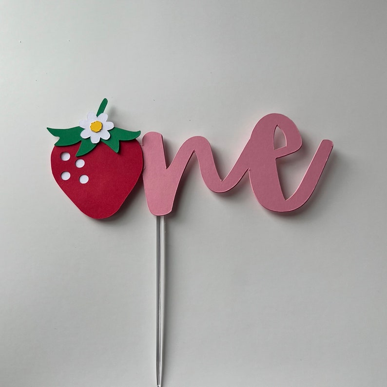 K&ouml;nnte beinhalten: Ein rosa Papier-Kuchentopper mit dem Wort "one" in kursivschrift. Eine rote Papier-Erdbeere mit gr&uuml;nen Bl&auml;ttern und einer wei&szlig;en Bl&uuml;te ist an der linken Seite des Toppers befestigt.