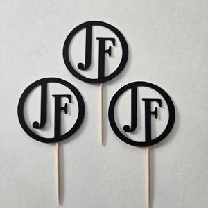 May include: Three black round cake toppers with the letters "JF" in white. Each topper is attached to a wooden stick.