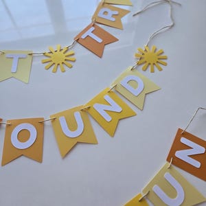 Puede incluir: Una pancarta de papel amarilla y naranja con las palabras "Round the Sun Trip" en letras blancas. La pancarta est&aacute; decorada con peque&ntilde;os soles de papel amarillo.