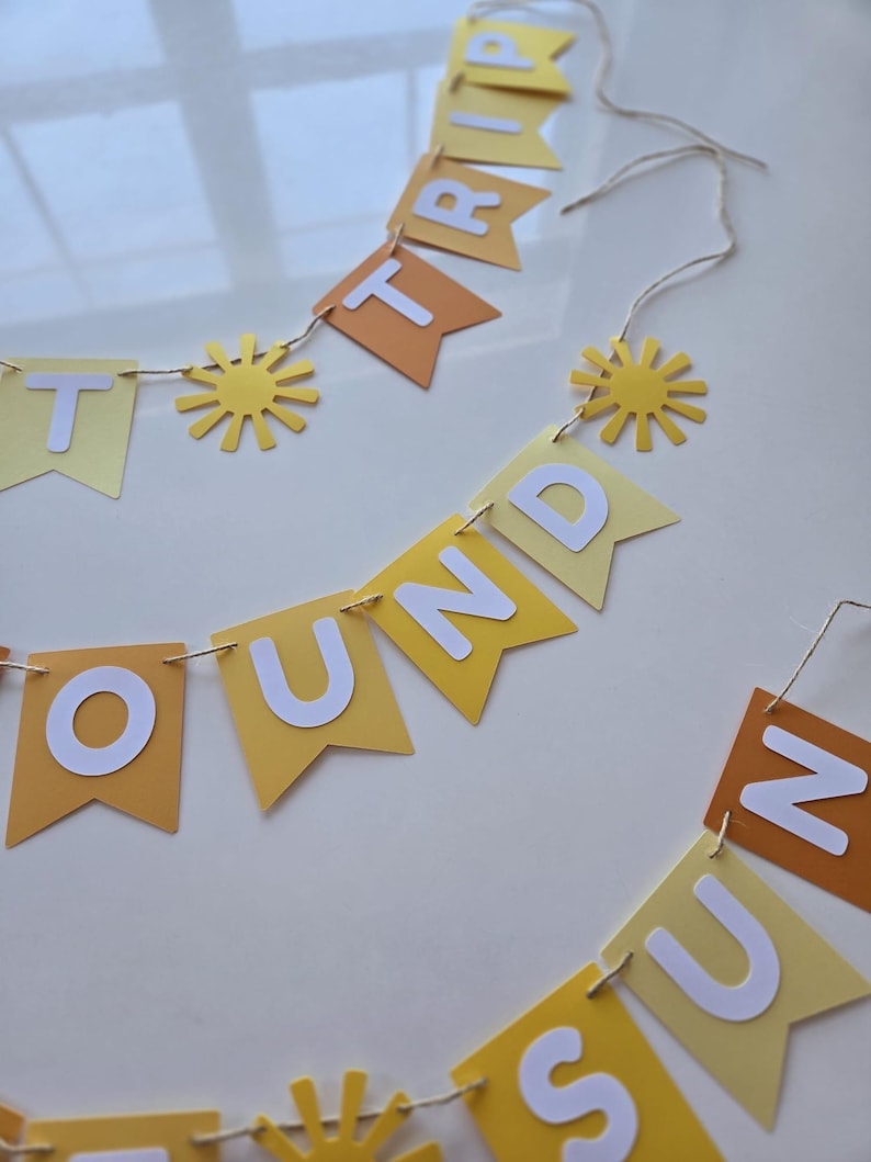 Puede incluir: Una pancarta de papel amarilla y naranja con las palabras "Round the Sun Trip" en letras blancas. La pancarta est&aacute; decorada con peque&ntilde;os soles de papel amarillo.