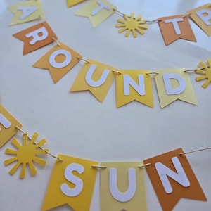 Puede incluir: Una pancarta de papel amarilla y naranja con las palabras "First Trip Around The Sun" y recortes de sol.