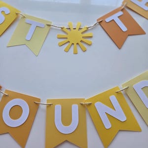 Puede incluir: Una pancarta de papel amarilla y naranja con las palabras "Just Around The" en letras blancas. La pancarta est&aacute; enhebrada con un cordel y tiene un sol de papel amarillo en el medio.