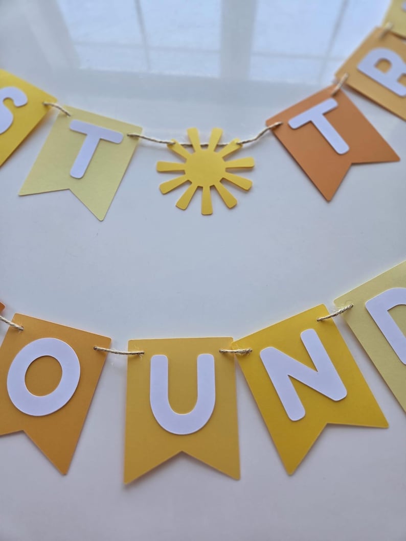 Puede incluir: Una pancarta de papel amarilla y naranja con las palabras "Just Around The" en letras blancas. La pancarta est&aacute; enhebrada con un cordel y tiene un sol de papel amarillo en el medio.
