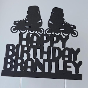 May include: Black cake topper featuring two roller skates above the words "HAPPY BIRTHDAY BRANTLEY". The text and skates are cut from black cardstock. The topper is attached to two thin white sticks.