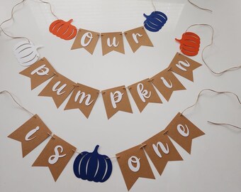 May include: A brown paper banner with white lettering that reads "Our Pumpkin is One". The banner is decorated with orange, white, and blue paper pumpkins.