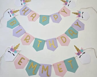 May include: A unicorn-themed birthday banner with the words "Happy Birthday Emmet" in gold glitter letters. The banner is made of pink, purple, and green paper with unicorn heads at each end.