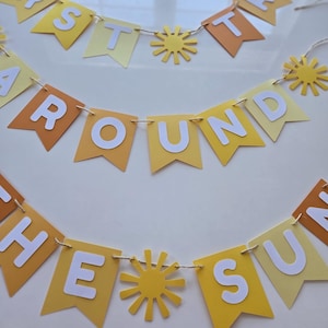 Puede incluir: Una pancarta de papel amarilla y naranja con las palabras "First Trip Around The Sun" en letras blancas. La pancarta est&aacute; decorada con soles de papel amarillo.