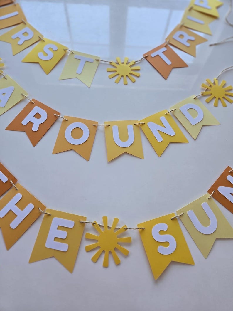 Puede incluir: Una pancarta de papel amarilla y naranja con las palabras "First Trip Around The Sun" en letras blancas. La pancarta est&aacute; decorada con soles de papel amarillo.