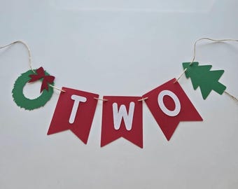 Op de afbeelding: Rode en groene kerstbanner met het woord "TWEE" in witte letters. De banner is versierd met een groene krans en een groene kerstboom.