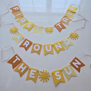 Puede incluir: Tres pancartas de papel amarillo y naranja con letras blancas que deletrean "First Trip Around The Sun". Cada pancarta tiene un peque&ntilde;o recorte de sol amarillo entre las palabras.