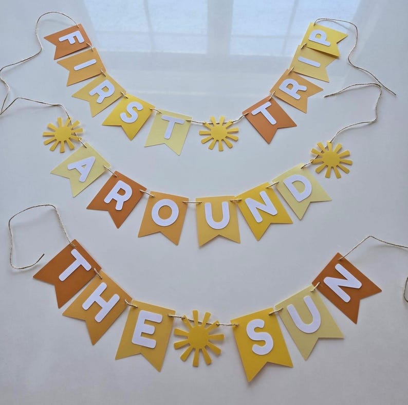 Puede incluir: Tres pancartas de papel amarillo y naranja con letras blancas que deletrean "First Trip Around The Sun". Cada pancarta tiene un peque&ntilde;o recorte de sol amarillo entre las palabras.