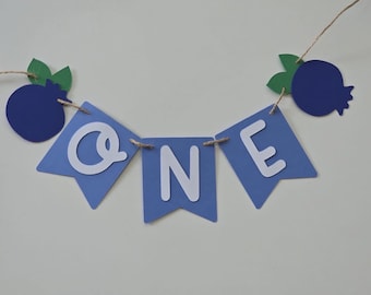 May include: A decorative banner featuring three light blue swallowtail pennants with the white letters O, N, and E. The banner is strung on twine and includes two dark blue blueberry-shaped cutouts with green leaves at each end of the string.