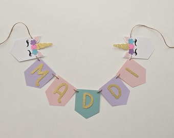 May include: A unicorn-themed banner with the name "MADDI" spelled out in gold glitter letters on pastel pink, purple, and green pennants. The banner is strung with twine and features two white unicorn heads with glitter horns and pink, purple, and blue flower accents.