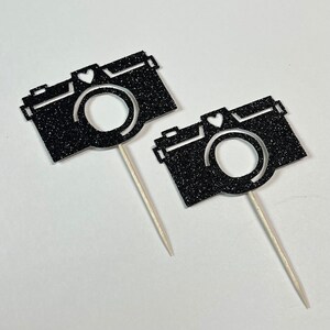 May include: Two black glitter camera cupcake toppers with white heart accents on wooden sticks.
