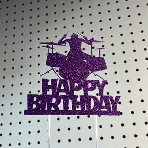 May include: Purple glitter cake topper featuring a drummer silhouette playing a drum set. The words "HAPPY BIRTHDAY" are below the drummer. The topper is mounted on a clear acrylic stick.