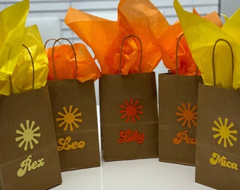 May include: Five brown paper gift bags with yellow and orange tissue paper. Each bag has a sun graphic and a name written in a retro font: Rex, Leo, Lily, Pat, and Mica.