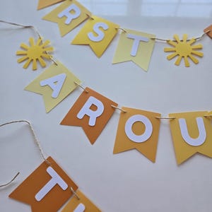 Puede incluir: Una pancarta de papel amarilla y naranja con las palabras "First Around The Sun" en letras blancas. La pancarta est&aacute; decorada con peque&ntilde;os soles de papel amarillo.
