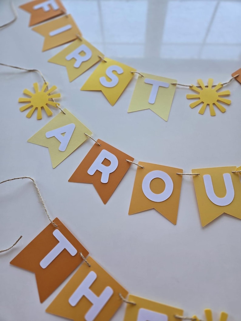 Puede incluir: Una pancarta de papel amarilla y naranja con las palabras "First Around The Sun" en letras blancas. La pancarta est&aacute; decorada con peque&ntilde;os soles de papel amarillo.